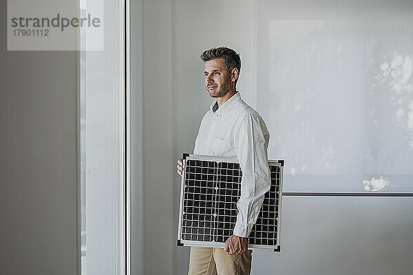 Lächelnder Ingenieur mit Solarpanel und Blick durch das Fenster im Büro