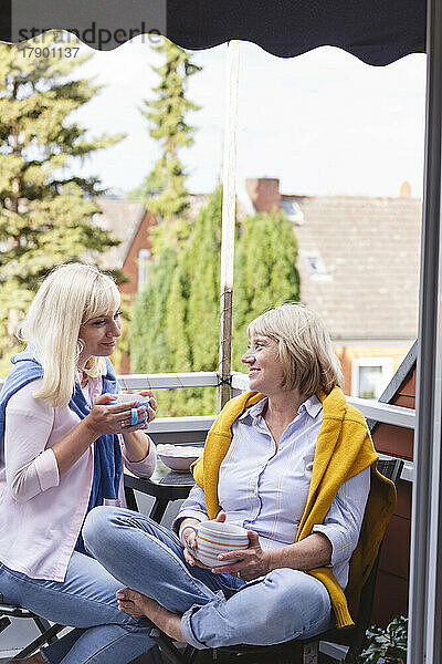 Lächelnde Mutter und Tochter mit Kaffeetassen sitzen zusammen auf dem Balkon