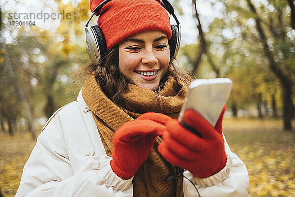 Glückliche junge Frau  die im Park Musik über Kopfhörer hört und ihr Smartphone nutzt