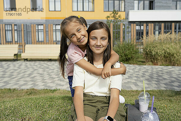 Glückliches Mädchen umarmt Schwester  die auf Gras im Park sitzt