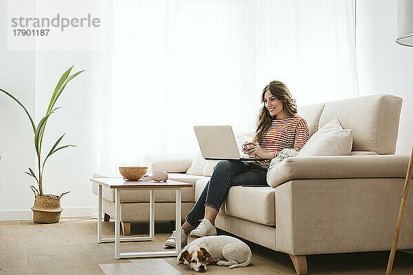 Junge Frau mit Hund benutzt Laptop auf Sofa zu Hause