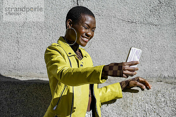 Glückliche Frau  die an einem sonnigen Tag ein Selfie mit dem Handy vor einer Betonwand macht