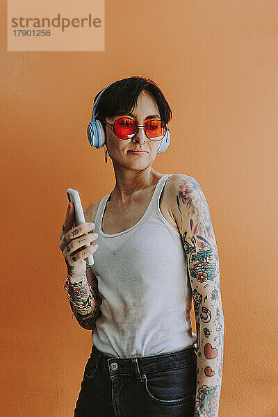 Beautiful woman listening music through wireless headphones standing against orange background