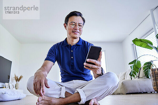 Happy mature man surfing net through mobile phone on floor at home
