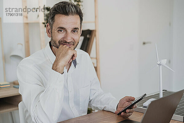 Lächelnder Geschäftsmann mit Smartphone und Laptop am Schreibtisch
