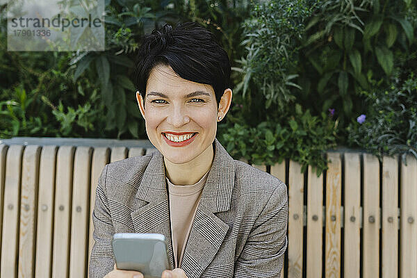 Glückliche Geschäftsfrau mit Smartphone sitzt auf Bank