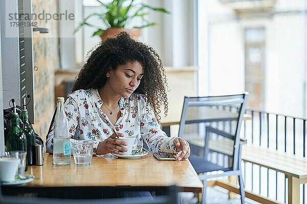 Junge Frau mit Kaffeetasse schaut im Café auf ihr Smartphone