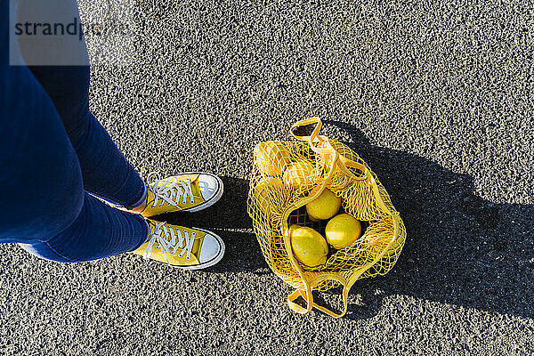 Frau steht auf Asphalt neben einem Netzbeutel mit Zitronen