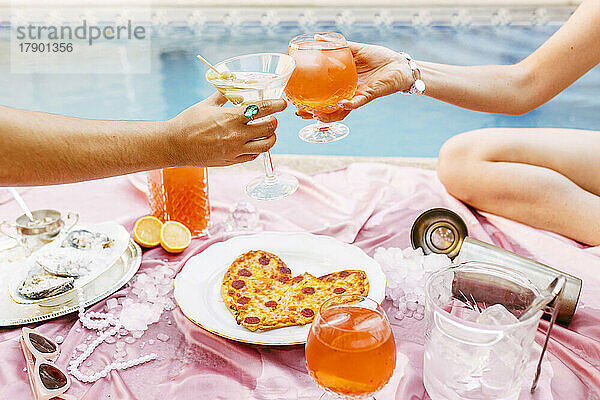 Freunde stoßen Cocktails bei herzförmiger Pizza am Pool an