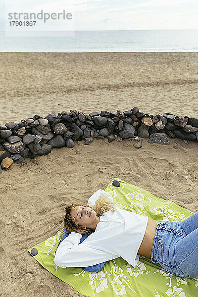 Entspannte Frau mit den Händen hinter dem Kopf liegt auf einer Picknickdecke am Strand