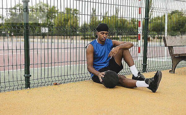 Junger Mann mit Basketball lehnt am Zaun und sitzt am Sportplatz