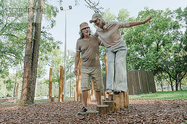 Älterer Mann unterstützt Frau beim Balancieren auf Baumstumpf im Park