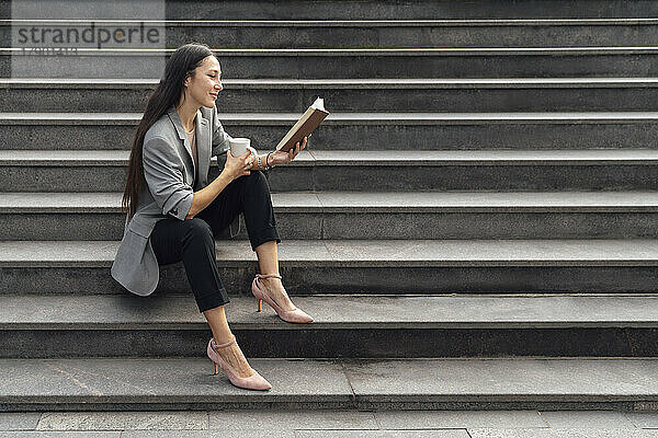 Lächelnde Geschäftsfrau liest Buch und sitzt mit Kaffeetasse auf der Treppe