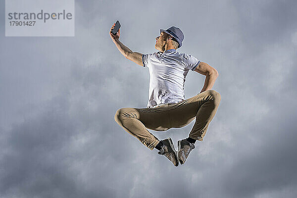 Mann springt und macht Selfie mit Smartphone unter bewölktem Himmel