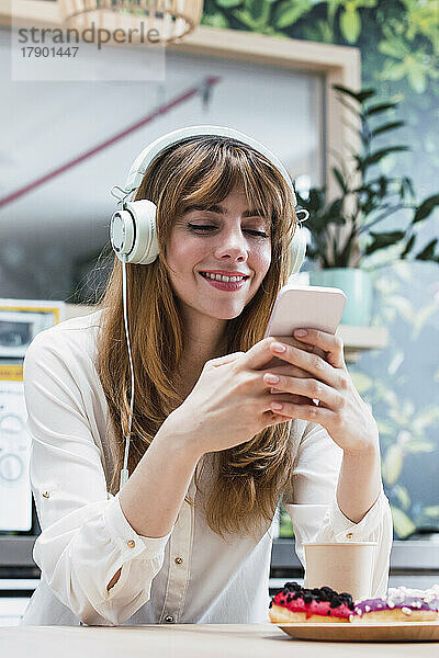 Lächelnde Geschäftsfrau mit Pony  die in der Cafeteria Textnachrichten über ihr Smartphone sendet