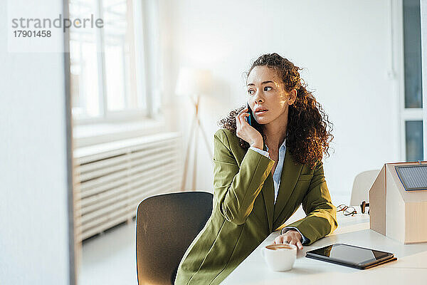 Junge Geschäftsfrau spricht mit Smartphone und sitzt mit Musterhaus am Schreibtisch im Büro