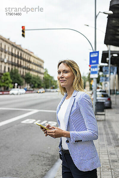 Lächelnde Geschäftsfrau mit Smartphone wartet am Bürgersteig auf Fahrt