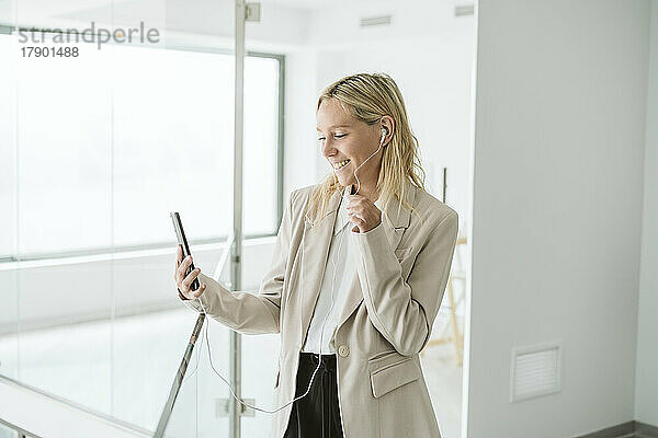 Lächelnde Geschäftsfrau blickt im Büroflur auf ihr Mobiltelefon
