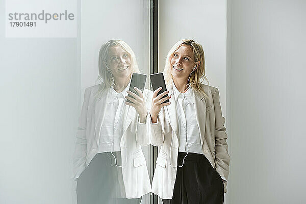 Lächelnde Geschäftsfrau hält Mobiltelefon an Glaswand