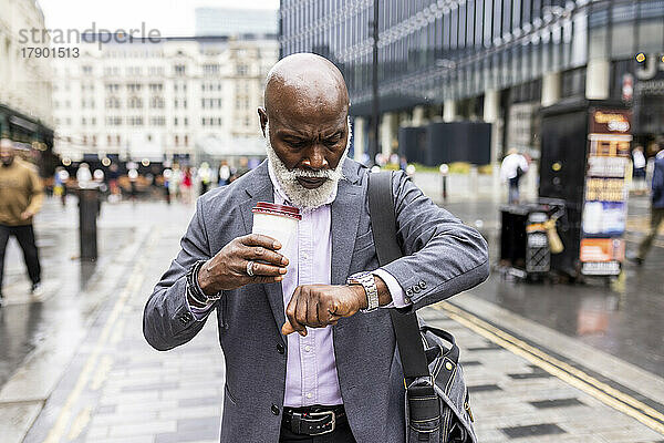 Leitender Geschäftsmann mit Einwegkaffeetasse  der in der Stadt die Uhrzeit auf der Armbanduhr überprüft