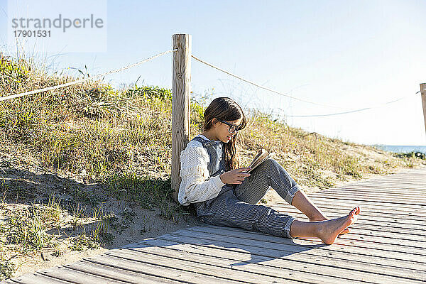 Mädchen liest Buch und lehnt an einer Stange an der Promenade