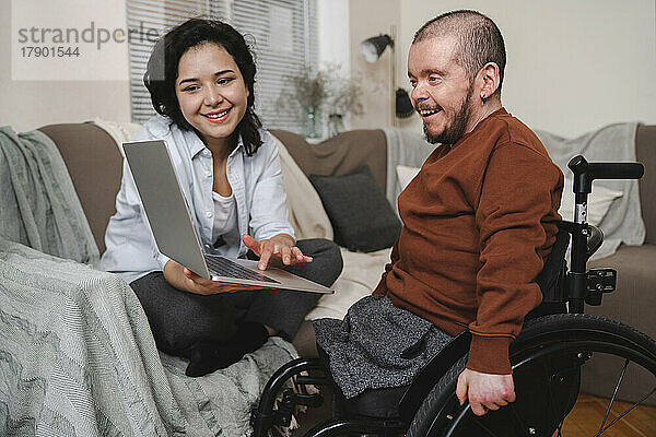 Lächelnde Frau zeigt ihrem Freund im Rollstuhl zu Hause Laptop