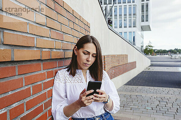 Ernsthafte Frau schaut auf ihr Mobiltelefon und lehnt an einer Ziegelwand