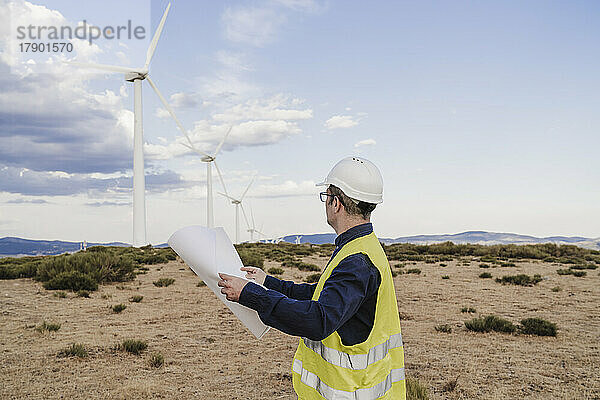 Ingenieur hält einen Bauplan in der Hand und betrachtet Windkraftanlagen im Windpark