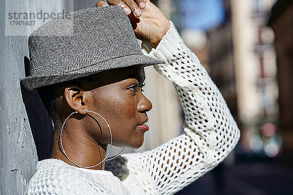 Young woman wearing hoop earrings and hat by wall on sunny day