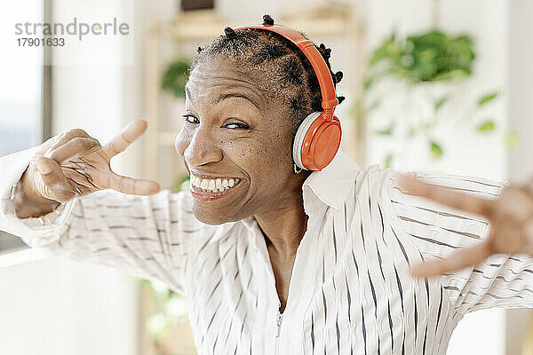 Glückliche Frau mit kabellosen Kopfhörern zeigt zu Hause ein Friedenszeichen