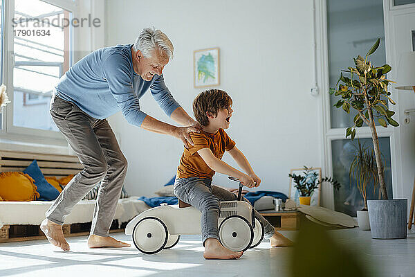 Verspielter älterer Mann schiebt seinen Enkel  der zu Hause auf einem Spielzeugauto sitzt