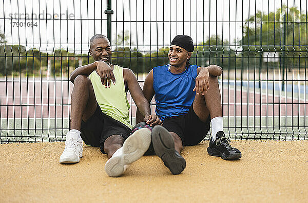 Lächelnder Sohn und Vater lehnen sich an den Zaun und sitzen auf dem Basketballplatz