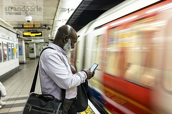 Glatzköpfiger älterer Geschäftsmann benutzt Smartphone in der Nähe eines rasenden Zuges am Bahnhof