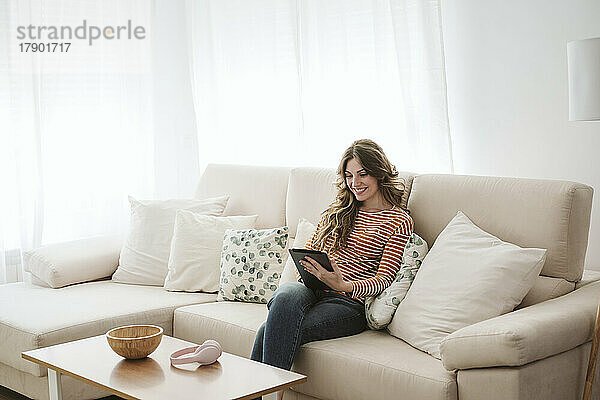 Lächelnde junge Frau nutzt digitales Tablet auf dem heimischen Sofa