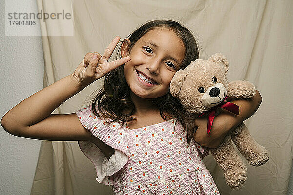 Happy girl with teddy bear gesturing peace sign
