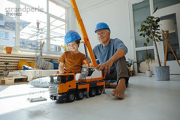 Lächelnder Enkel und Großvater in Schutzhelmen mit Spielzeugkran und Strommastmodell zu Hause