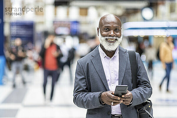 Lächelnder kahlköpfiger Pendler mit Mobiltelefon am Bahnhof