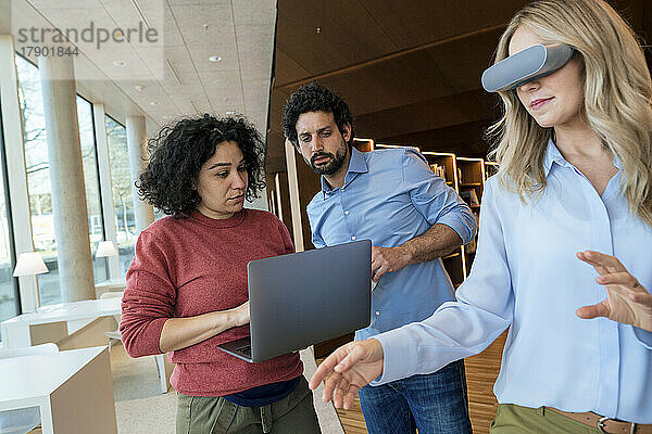 Frau mit VR-Brille von Kollegen mit Laptop in der Bibliothek