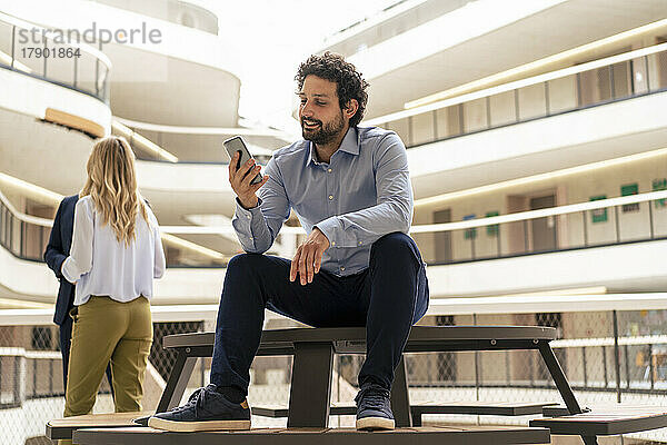 Lächelnder Geschäftsmann nutzt Smartphone mit Kollegen im Hintergrund am Flur