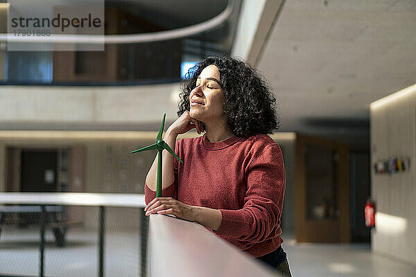 Geschäftsfrau mit geschlossenen Augen hält Windkraftanlage am Geländer im Bürokorridor