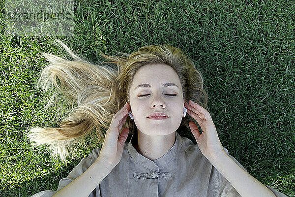 Woman with blond hair listening music through wireless in-ear headphones lying on grass