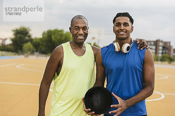 Lächelnder junger Mann hält Basketball und steht mit seinem Vater auf dem Sportplatz