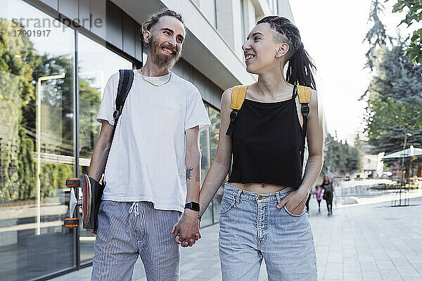 Glückliches junges Hipster-Paar hält sich an den Händen und geht auf dem Fußweg