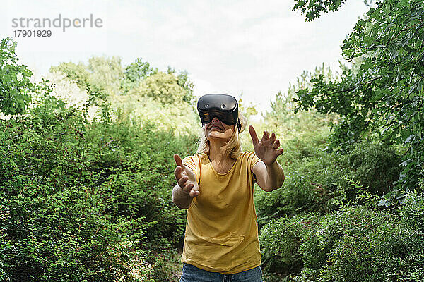 Reife Frau gestikuliert mit Virtual-Reality-Simulatoren  die inmitten von Pflanzen stehen