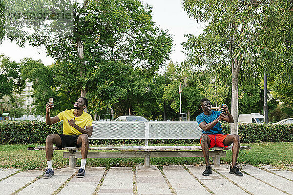 Junge Männer machen Selfies mit Smartphones und sitzen auf einer Bank im Park