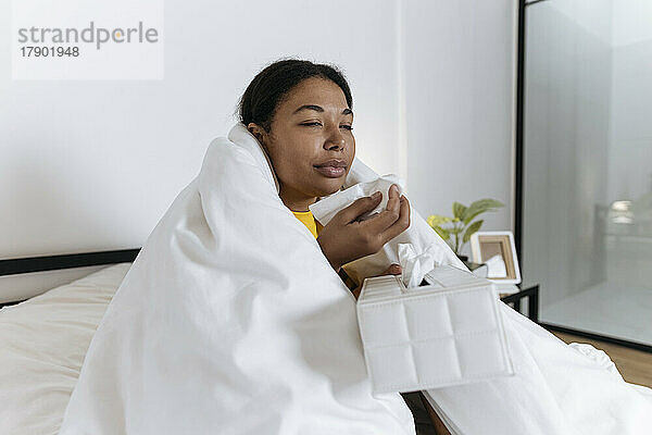 Kranke Frau sitzt auf dem Bett  in eine Decke gehüllt und putzt sich die Nase in ein Taschentuch