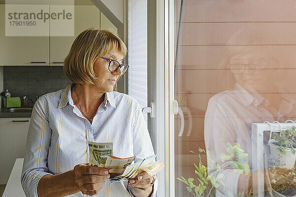 Ältere Frau mit Banknoten  die zu Hause durch das Fenster schaut