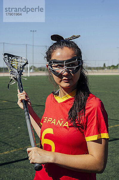 Nachdenklicher junger Spieler mit Lacrosse-Schläger und Schutzbrille auf dem Sportplatz
