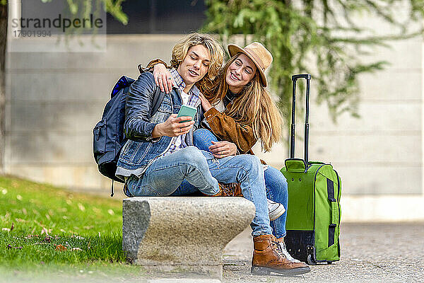 Mann teilt Smartphone mit Frau  die neben Gepäck auf Bank sitzt