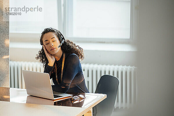 Junge Geschäftsfrau mit Headset sitzt mit geschlossenen Augen am Schreibtisch im Büro